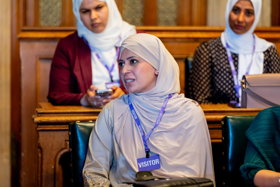 Kanza Azher listens attentively during the APPG on Hajj & Umrah meeting at the Palace of Westminster, wearing a visitor pass and seated among attendees in the public gallery.